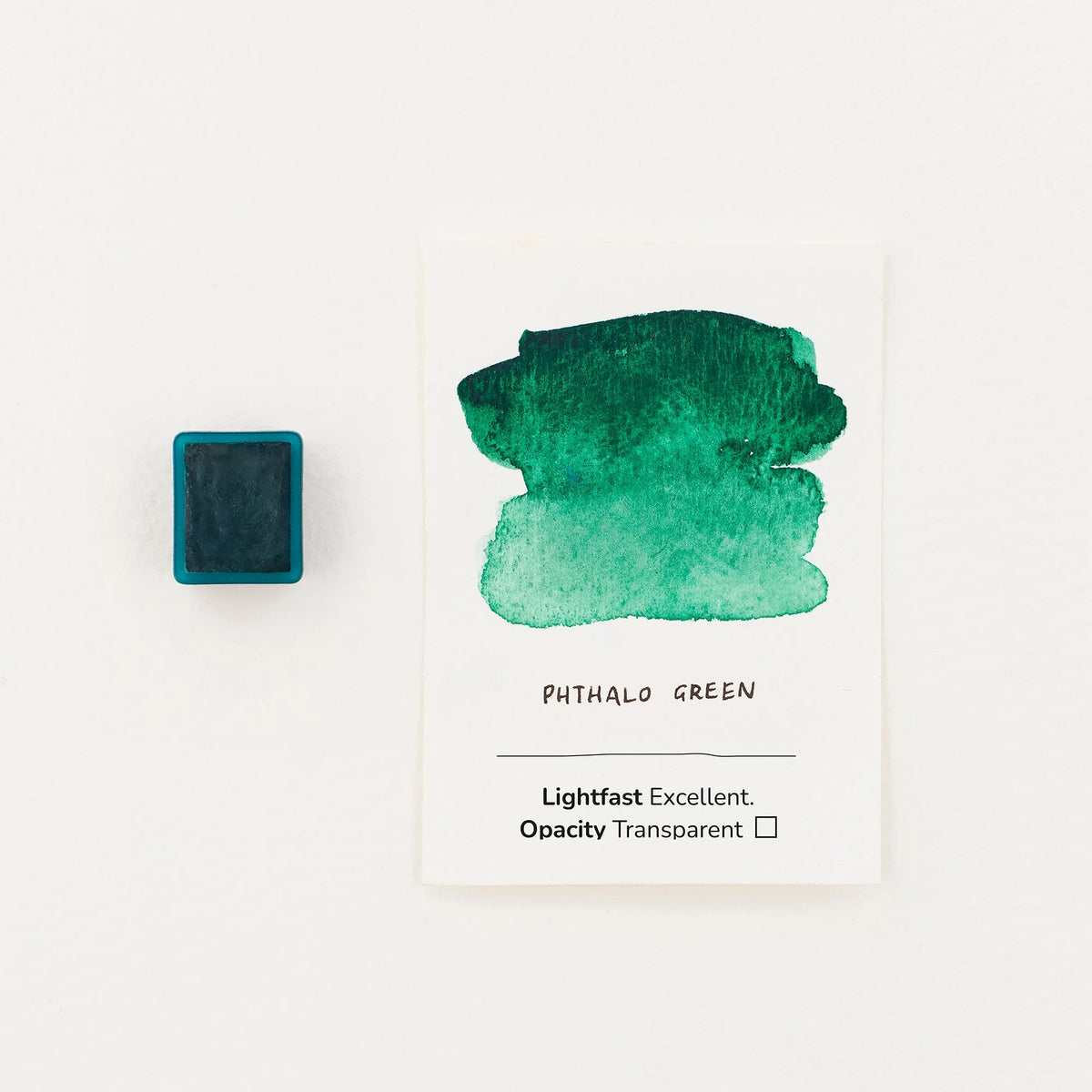 A watercolor paint swatch card labeled 'PHTHALO GREEN' with a green wash on white paper, shown next to a watercolour half pan on a clean white background.