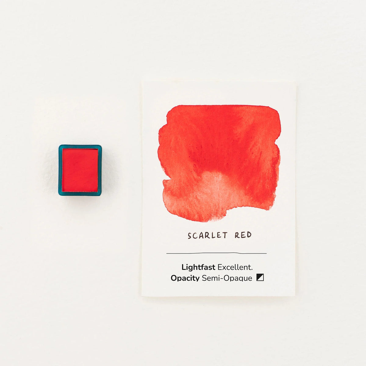 A square watercolour half pan in a teal case sits to the left of a white card displaying a watercolor sample of scarlet red, with text reading 'SCARLET RED' and 'Lightfast Excellent, Opacity Semi-Opaque'.