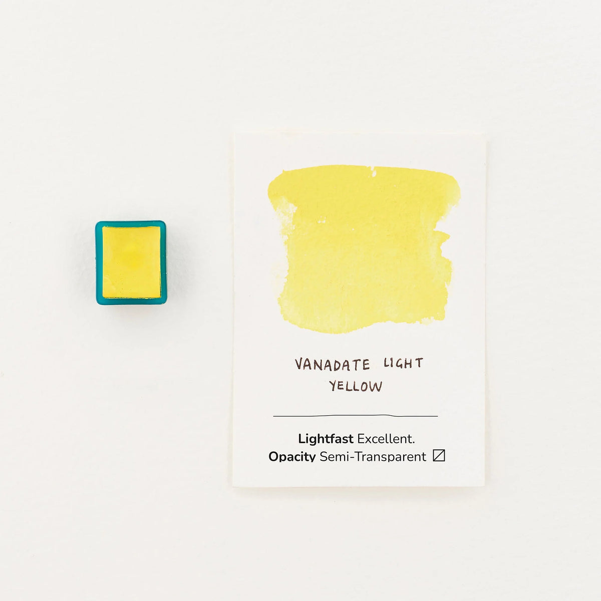A square pigment cube with a yellow center and teal-blue border sits to the left of a white card labeled 'VANADATE LIGHT YELLOW' with a painted yellow swatch above it, on a clean white background.