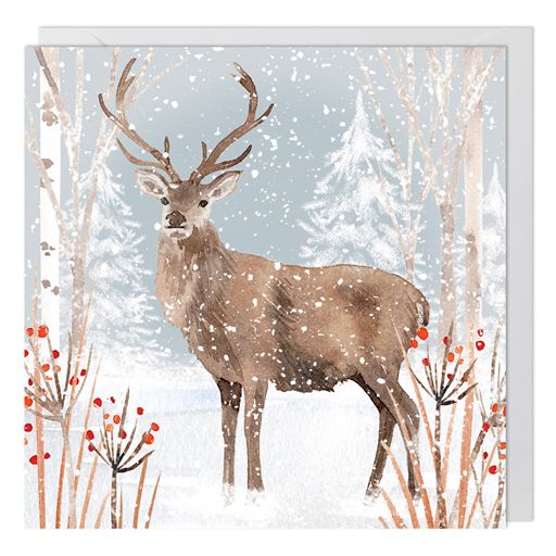 A square greeting card featuring a watercolor-style brown stag with antlers standing in a snowy winter forest, with birch trees, falling snow, and small red berries. A white envelope is partially visible behind the card.