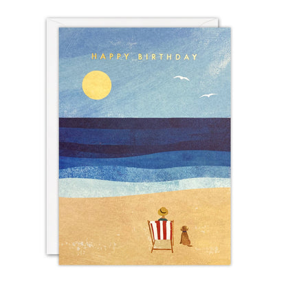 Birthday card of a person in a striped deckchair with a dog on a sandy beach under a yellow sun, gold foil text