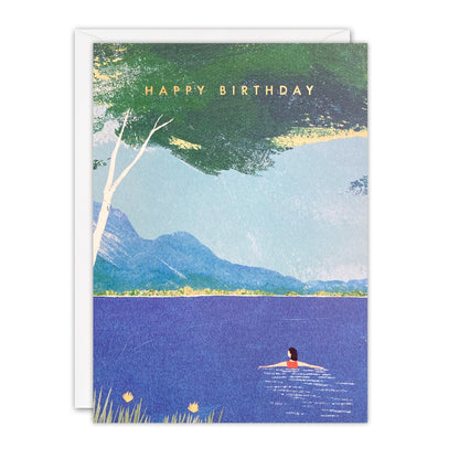 Birthday card of a woman wild swimming in a deep blue lake below mountains and a birch tree, gold foil text
