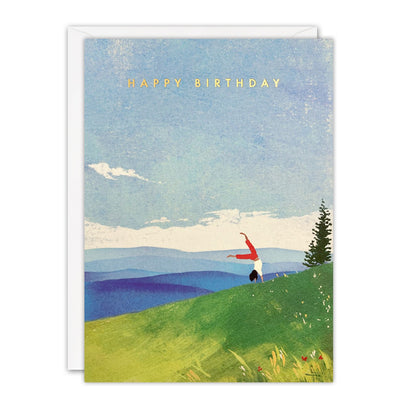 Birthday card of a person cartwheeling on a green hillside above blue hills and a pine tree, gold foil text